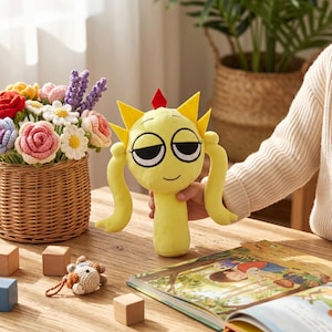 Puede incluir: Un peluche amarillo con una cara sonriente, ojos negros y pelo amarillo puntiagudo es sostenido. El juguete tiene brazos largos y curvos. Una cesta de flores de ganchillo coloridas y un libro infantil están en el fondo.