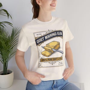 May include: Cream-colored t-shirt featuring a graphic with the text "Quiet Mornings Club" and an illustration of butter being sliced. The design includes the words "Salted" and "100% Pure Soft Life".