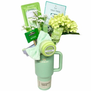 Op de afbeelding: Een mintgroene Stanley Quencher H2.0 Tumbler bevat een verzameling groene items. Deze omvatten een gezichtsmasker, lippenbalsem, Ghirardelli mintkoekje en een klein boeket witte bloemen en groene hortensia's. Een groene strik en handontsmettingsmiddel zijn ook inbegrepen.