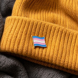 Könnte beinhalten: Eine senfgelbe, gerippte Strickmütze mit einer kleinen Emaille-Anstecknadel in Form einer wehenden Flagge. Die Flaggennadel zeigt horizontale Streifen in Hellblau, Rosa und Weiß, die die Farben der Transgender-Flagge mit einem Goldrand darstellen.