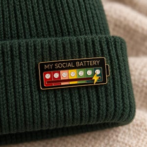 Könnte beinhalten: Eine dunkelgrüne, gerippte Strickmütze mit einer rechteckigen Emaille-Anstecknadel. Die Nadel zeigt eine Batteriestandsanzeige mit dem Text MY SOCIAL BATTERY, einem Farbverlauf von Rot nach Grün, verschiedenen Gesichtern und einem gelben Blitz.