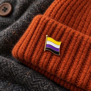 Könnte beinhalten: Eine kleine Metall-Emaille-Anstecknadel in Form einer wehenden Flagge mit horizontalen Streifen in Gelb, Weiß, Violett und Schwarz. Die Anstecknadel ist an einer dicken, gerippten Strickmütze in Rostorange befestigt, die auf einem dunkelgrauen Wollstoff liegt.