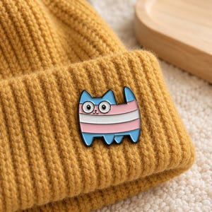 Könnte beinhalten: Eine senfgelbe, gerippte Strickmütze mit einer Metall-Emaille-Anstecknadel in Form einer Katze. Die Katzen-Anstecknadel hat eine schwarze Umrandung und ist mit horizontalen Streifen in Hellblau, Rosa und Weiß sowie zwei großen weißen Augen versehen.