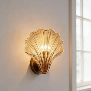 Puede incluir: Lámpara de pared decorativa con forma de concha. La lámpara es de vidrio color ámbar con textura acanalada, montada sobre una base circular de latón. La luz brilla a través del vidrio, creando un brillo cálido. Montada en una pared blanca.