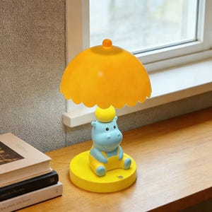 May include: A novelty lamp in the shape of a hippo, with a yellow lampshade and base, and a blue hippo body wearing yellow overalls. The lamp is on a wooden surface. The lampshade has a scalloped edge.