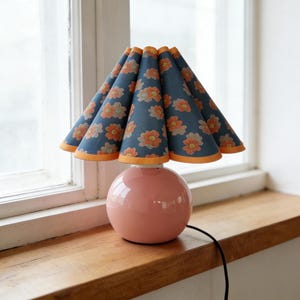May include: A pink ceramic table lamp with a round base and a navy blue lampshade. The lampshade has an orange and peach floral pattern. The lamp is on a wooden surface near a window.