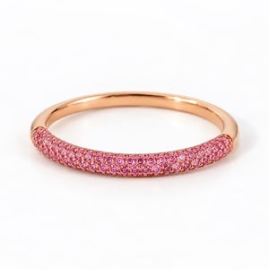 May include: A rose gold band ring featuring a pavé setting of numerous small, vibrant pink stones covering the front half of the circular band. The metal has a smooth, polished finish, and the pink stones are arranged in a dense, uniform pattern.