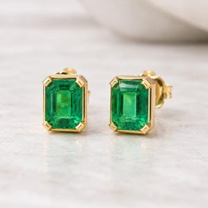 Könnte beinhalten: Ein Paar Ohrstecker mit rechteckigen, smaragdgeschliffenen grünen Edelsteinen, die in polierte Gelbgoldfassungen mit vier Eckkrappen eingefasst sind. Die Ohrringe haben ein klassisches Design mit einem leuchtend grünen Farbton.