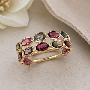 May include: A gold ring featuring a double row of oval-cut gemstones in shades of pink, purple, green, and blue. The ring's band is a polished gold, and the gemstones are set closely together.