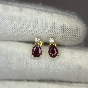 Może przedstawiać: Para złotych kolczyków z gruszkowymi, głęboko czerwonymi kamieniami szlachetnymi. Każdy kolczyk ma mały, okrągły, przezroczysty kamień na górze. Kolczyki są na białym tle.