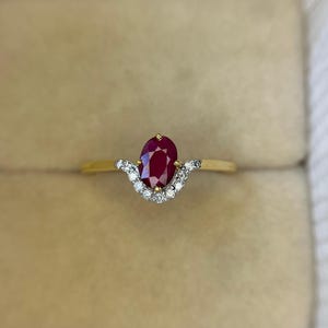 Può includere: Un anello d'oro con una grande gemma di rubino ovale. Il rubino è di un colore rosso intenso ed è circondato da una fila di piccoli diamanti trasparenti. L'anello ha una sottile fascia d'oro.