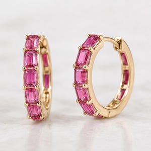 May include: A pair of gold hoop earrings featuring a row of vibrant pink emerald-cut gemstones set along the front. Each hoop is crafted with a polished gold metal finish and a secure hinged closure, showcasing a series of five rectangular pink stones.