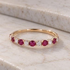 May include: A gold band ring featuring an alternating pattern of round-cut red rubies and clear white diamonds set in a shared prong setting. The ring has a polished metal finish and displays a continuous row of gemstones across the front half.