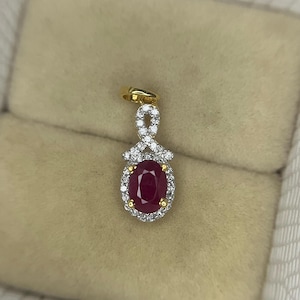 Könnte beinhalten: Goldener Anhänger mit einem ovalen Rubin, umgeben von kleinen Diamanten. Der Anhänger hat ein aufwendiges Design und eine Schlaufe für eine Kette. Der Rubin hat eine tiefe rote Farbe. Präsentiert auf beigem Untergrund.