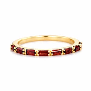 Puede incluir: Un anillo de banda dorado con una serie de piedras preciosas rectangulares de color rojo intenso. El anillo tiene una superficie pulida y reflectante, y las piedras preciosas están espaciadas uniformemente alrededor de la banda. El anillo está sobre un fondo blanco.