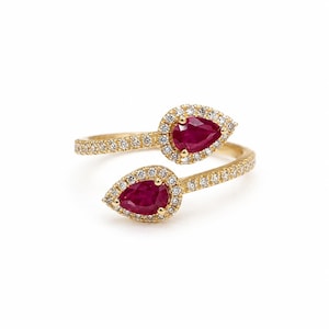 May include: A bypass ring crafted from yellow gold, featuring two pear-shaped red rubies at the terminals. Each ruby is surrounded by a halo of small white diamonds, and the band is pavé-set with additional white diamonds along the shank.