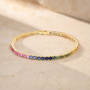 May include: A 14K gold tennis bracelet featuring a continuous row of round-cut gemstones in a gradient of colours, transitioning from pink and purple to blue, green, and yellow. The metal clasp is clearly engraved with the text 14K.