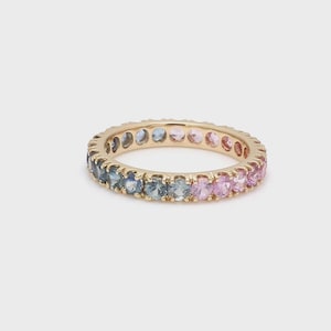 May include: A gold eternity ring featuring a continuous row of alternating blue and pink gemstones. The ring's design showcases a full circle of colourful stones. The ring is displayed on a plain, light background.