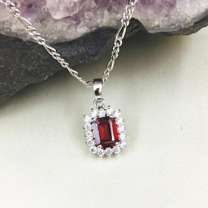 May include: A silver necklace with a rectangular, deep red pendant surrounded by small, clear stones. The pendant hangs from a delicate chain, showcasing a classic and elegant design. The necklace is set against a neutral background.
