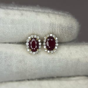 May include: A pair of stud earrings featuring oval-cut, deep red gemstones surrounded by a halo of smaller, clear stones. The earrings are set in a gold-colored metal, creating a classic and elegant design. The gemstones are the focal point.