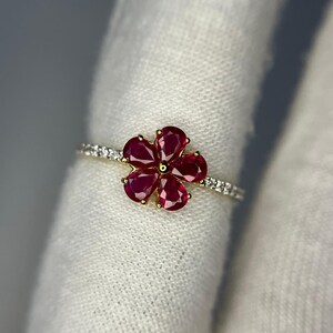 Puede incluir: Un anillo de oro con un diseño floral con cinco pétalos de piedras preciosas rojas. La banda está adornada con pequeñas piedras preciosas transparentes. El anillo está sobre un fondo blanco.