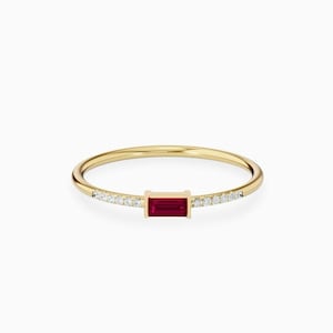 Peut inclure: Une fine bague en or avec une pierre rouge foncé rectangulaire au centre. La monture est fine, avec de petites pierres claires de chaque côté de la pierre centrale, créant un accent scintillant. La bague est posée sur un fond blanc.
