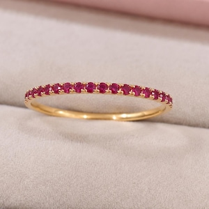 May include: A thin gold band ring featuring a continuous row of small, round, vibrant pinkish-red gemstones set along the top half of the metal circumference, creating a delicate and uniform circular design with a polished metallic finish.
