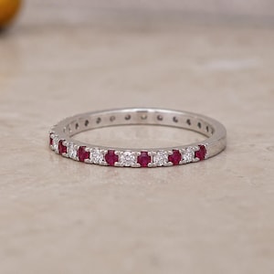 Puede incluir: Un anillo de banda fina en tono plateado con un patrón alterno de rubíes rojos redondos y piedras blancas transparentes engastadas a lo largo de la mitad frontal de la banda. El metal tiene un acabado pulido y las piedras están en fila.
