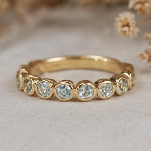 Puede incluir: Un anillo de oro con una serie de piedras preciosas redondas de color azul claro engastadas en biseles individuales. Las piedras están dispuestas en una fila continua a lo largo de la mitad superior de la banda, con un acabado metálico pulido.