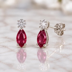 May include: A pair of silver-toned stud earrings featuring a marquise-cut deep red gemstone suspended beneath a small, round, clear faceted stone. The earrings have a post-back closure and are displayed with their reflections visible below.