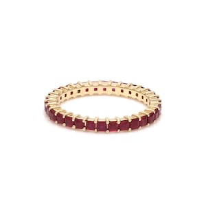 May include: A gold-colored eternity ring featuring a continuous row of square-cut, deep red gemstones. The ring is set against a white background, highlighting the vibrant color and design of the jewelry.