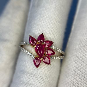 May include: A gold ring featuring a floral design with vibrant red ruby petals. The ring's band splits into two, each lined with small, sparkling white gemstones. The central ruby is surrounded by six pear-shaped rubies.