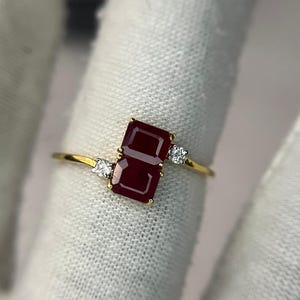 May include: A gold ring featuring two rectangular, deep red gemstones. Small, clear gemstones flank the larger stones. The ring's band is thin and gold. The ring is set against a white background.