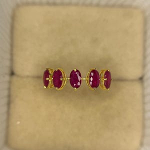 Puede incluir: Un anillo de oro con cinco piedras preciosas rojas oscuras de forma ovalada. Las piedras están engarzadas en una fila, con una delicada banda de oro. El anillo se presenta en un joyero beige, resaltando su diseño y color.