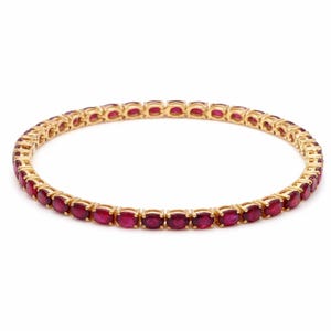 May include: A gold-toned bracelet featuring a continuous line of oval-cut, deep red gemstones. The gemstones are set closely together, creating a vibrant, eye-catching piece of jewelry. The bracelet is designed with a classic, elegant style.