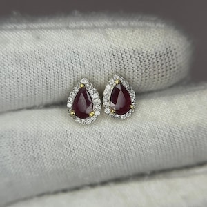 May include: A pair of teardrop-shaped earrings featuring deep red rubies at the center, each surrounded by a halo of small, clear, round-cut stones set in a metallic frame with yellow gold prongs securing the central red gemstones.