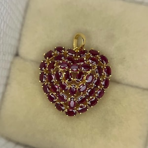 Op de afbeelding: Een hartvormige hanger met een gouden zetting, dicht bezet met ovaal geslepen, dieprode edelstenen. De hanger heeft een kleine lus aan de bovenkant voor een ketting. De edelstenen zijn zo gerangschikt dat ze een textuur creëren.