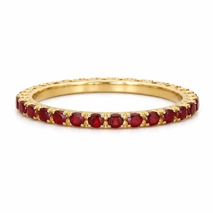 May include: A gold band ring featuring a series of small, round, bright red gemstones. The ring has a detailed pattern along the edges, and the gemstones are evenly spaced around the band. The ring is set against a white background.