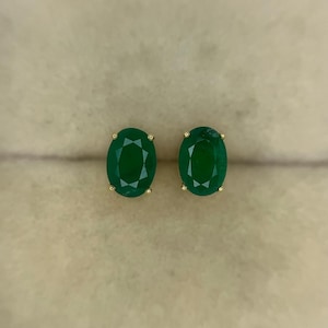 Puede incluir: Un par de pendientes de botón de esmeralda de corte ovalado. Las vibrantes piedras preciosas verdes están engastadas en garras de oro. Los pendientes se muestran sobre un fondo neutro.