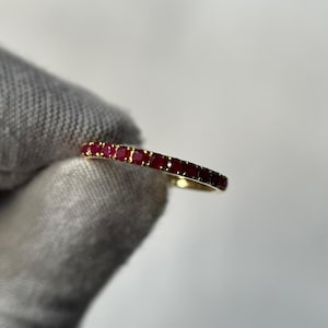 May include: A gold band ring set with a row of small, vibrant red gemstones. The ring is held against a neutral background, showcasing the ring's design and color.