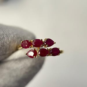 Può includere: Un anello d'oro con sette rubini ovali e a forma di pera. I rubini sono di un rosso intenso e sono incastonati in un design delicato e curvo. L'anello è mostrato su uno sfondo neutro.