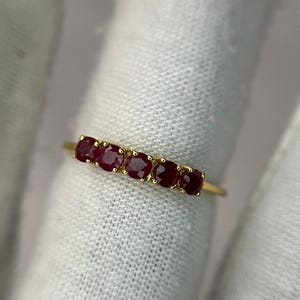May include: A gold ring featuring five oval-cut, deep red gemstones. The gemstones are set in a row along the top of the ring. The ring is set against a white background.