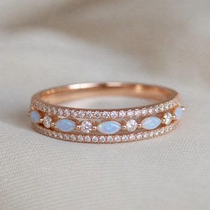 May include: Rose gold ring with three rows of embellishments. The top and bottom rows are encrusted with small, clear stones. The middle row features oval opals and round, clear stones. The ring is set against a soft, neutral background.
