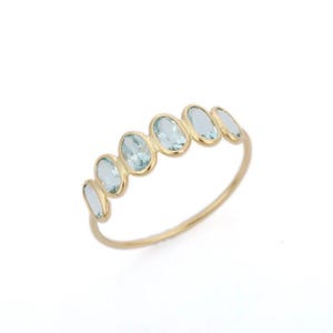 Op de afbeelding: Een gouden ring met zes ovale, lichtblauwe edelstenen. De edelstenen zijn in een rij op de ringband geplaatst. De ring heeft een dunne, delicate band. De edelstenen zijn gelijkmatig verdeeld. Witte achtergrond.