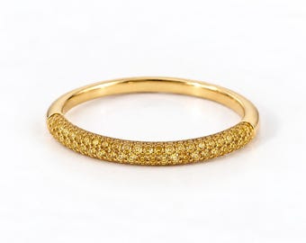 May include: A gold band ring featuring a pavé setting of numerous small, round, yellow-toned gemstones covering the front half of the circular band. The metal has a polished, reflective finish, and the stones are arranged in a dense, uniform pattern.