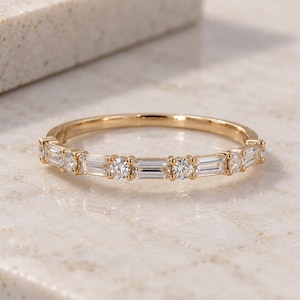 May include: A gold band ring featuring an alternating pattern of clear baguette-cut stones and round brilliant-cut stones set along the top half of the band. The metal has a polished finish, and the stones are arranged in a repeating sequence.