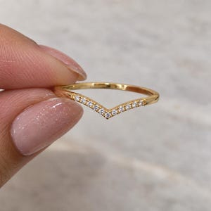 May include: A gold V-shaped chevron ring featuring a row of small, clear, round-cut stones set along the centre point of the band. The polished metal band has a smooth, thin profile that tapers slightly towards the back, creating a delicate appearance.