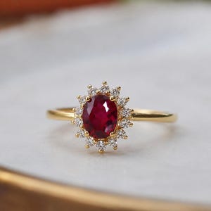 Può includere: Un anello in oro con un rubino centrale rosso intenso dal taglio ovale, circondato da un alone di piccoli diamanti bianchi rotondi e trasparenti. Il gambo è sottile e in oro lucido, creando un design classico e simmetrico dai colori vivaci.