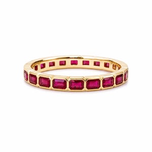 May include: A yellow gold eternity band featuring a continuous row of rectangular, step-cut vibrant red rubies set in a channel setting. The ring has a polished metallic finish and a uniform design that encircles the entire circumference of the band.