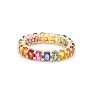 May include: A gold ring featuring a continuous row of oval-cut gemstones in a rainbow of colors. The gemstones include red, orange, yellow, green, blue, and purple hues, creating a vibrant and eye-catching design.